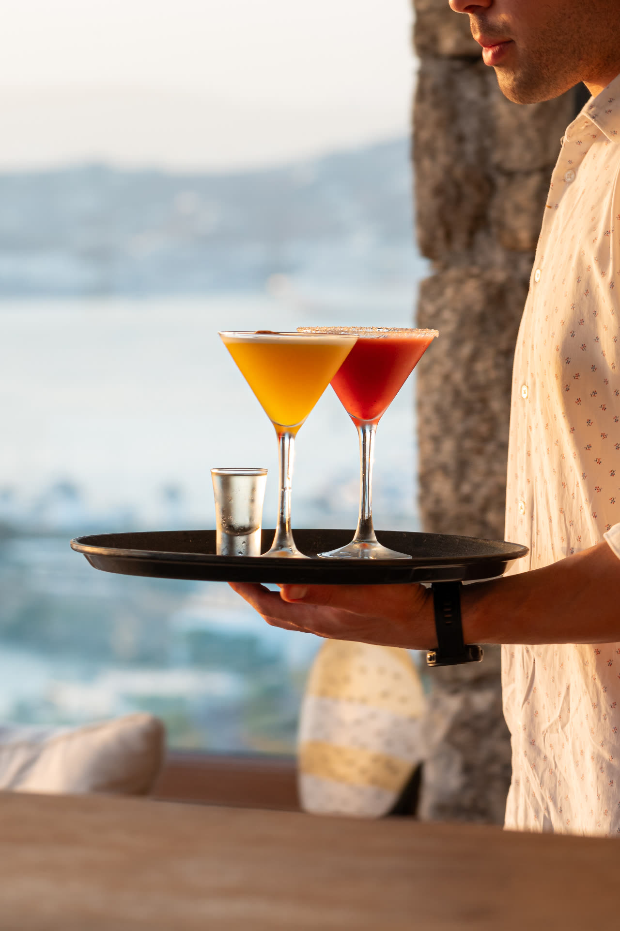 Server carrying a tray of signature sunset cocktails above Mykonos